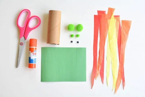 Scissors, glue stcik, toilet paper roll, green paper, findings for dragon face, orange and yellow strips of paper lined up in order of usage for craft