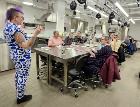 Woman in print jumpsuit stands in front of a table speaking to group of adults seated at a table in an indutrial kitchen